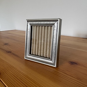 May include: A rectangular silver-toned picture frame with an ornate, engraved leaf pattern along the border. The frame contains a piece of light brown, vertically ribbed corrugated cardboard as a placeholder instead of a photograph.