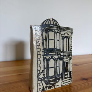 May include: A ceramic art piece depicting a building facade with a domed roof and detailed window designs. The artwork is primarily off-white with black ink detailing, showcasing a unique architectural illustration. The piece is displayed on a wooden surface.