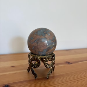 May include: A decorative sphere with a mottled blue and brown pattern, resting on an ornate brass-colored stand. The stand features a detailed, openwork design with curved elements and three small feet. The sphere appears to be made of stone or a similar material.