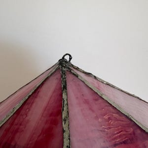 May include: A close-up of a stained glass lampshade with a pyramid shape. The glass is a gradient of pink and red hues, with a textured surface. The frame is a dark gray color.