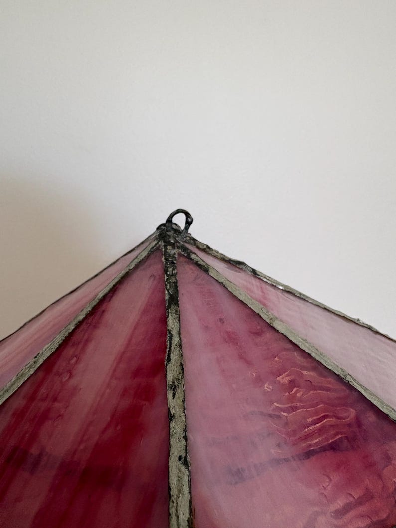 May include: A close-up of a stained glass lampshade with a pyramid shape. The glass is a gradient of pink and red hues, with a textured surface. The frame is a dark gray color.