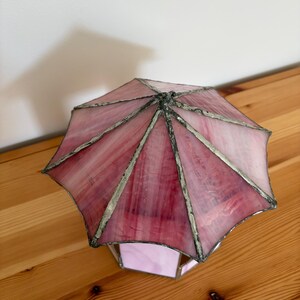 May include: A stained glass lamp with a pink and white umbrella-shaped shade. The shade is made of multiple glass panels, each with a different shade of pink. The lamp has a silver-colored metal frame and a geometric base.