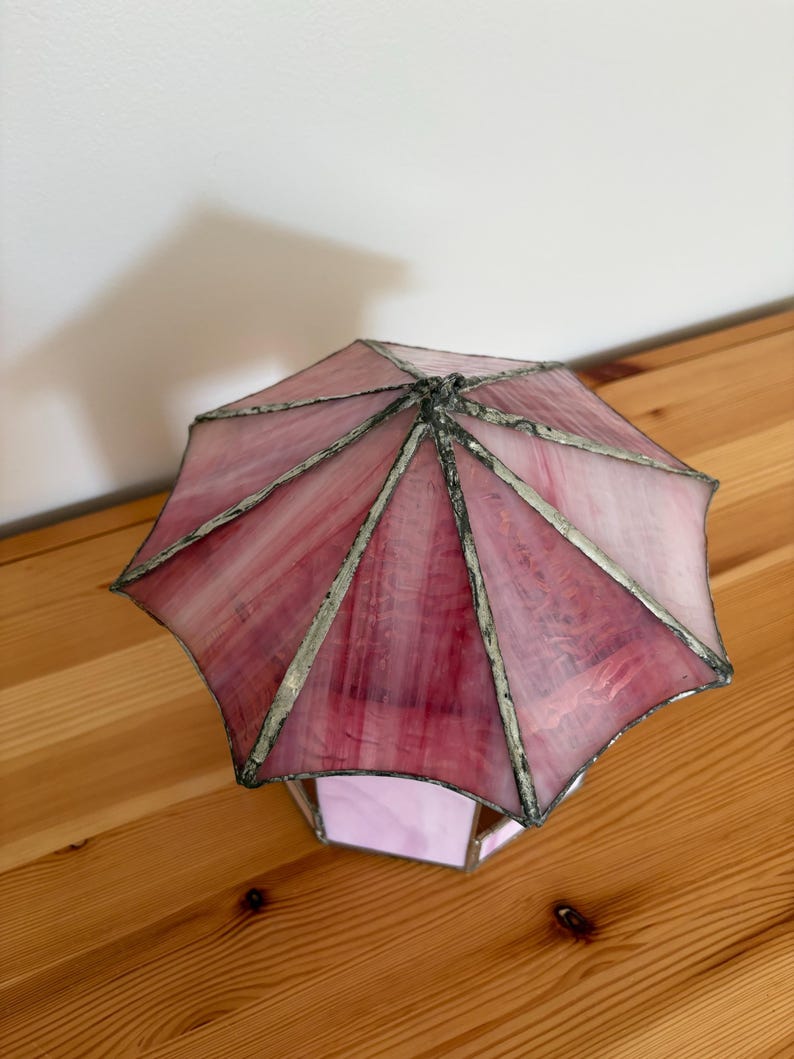 May include: A stained glass lamp with a pink and white umbrella-shaped shade. The shade is made of multiple glass panels, each with a different shade of pink. The lamp has a silver-colored metal frame and a geometric base.
