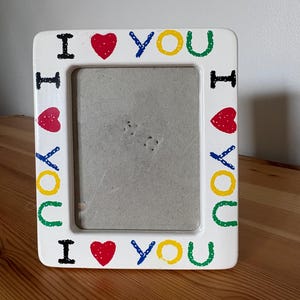 May include: A rectangular white picture frame featuring the text I LOVE YOU repeated around the border. The letters are painted in black, yellow, and green, with a red heart symbol replacing the word LOVE in each instance of the phrase.