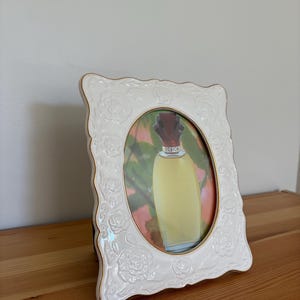 May include: White oval picture frame with a floral design and gold trim. The frame holds a picture of a perfume bottle with the word "DESIGN" visible on the bottle.