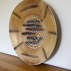 Puede incluir: Un plato redondo de cerámica con fondo beige y centro texturizado. El centro presenta detalles moteados en blanco y negro con líneas negras horizontales. Líneas marrones oscuras irradian desde el centro.