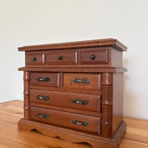 May include: A wooden jewelry box with multiple drawers and a top compartment. The box is a warm brown color with dark hardware and decorative accents. The box has a rectangular shape and is made of wood.