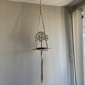 May include: A macrame plant hanger with a wooden shelf and a black metal tree-shaped jewelry holder. The hanger is made of natural-colored rope and has a tassel at the bottom.
