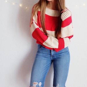 Può includere: Un maglione a righe rosse e bianche con maniche lunghe e un taglio ampio. Il maglione è realizzato in un tessuto morbido e lavorato a maglia.