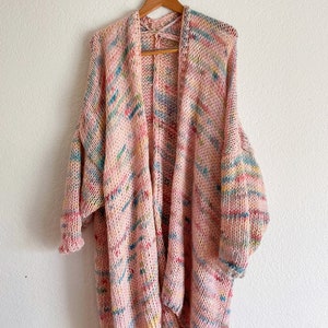 Op de afbeelding: Een lange, roze cardigan met een veelkleurig garenpatroon. De cardigan heeft een losse pasvorm en lange mouwen. Hij hangt aan een houten hanger.