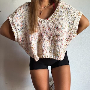 May include: A cream-colored, short-sleeved, hand-knitted sweater featuring a speckled pattern with small flecks of blue, yellow, pink, and orange throughout the yarn. The garment has a wide, rounded neckline and a loose, boxy, cropped fit.
