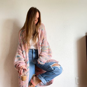 Op de afbeelding: Een vrouw draagt een roze en wit gestreepte cardigan met een wit tanktop en blauwe jeans. Ze houdt een kop koffie vast en heeft haar voet op een stoel.