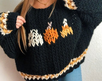 May include: Black oversized knit sweater with a white, orange, and yellow candy corn design. The sweater has a ribbed trim around the neckline, cuffs, and hem.