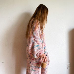 Op de afbeelding: Een lange, roze cardigan met een kleurrijk, gevlekt patroon. De cardigan is gebreid met een los, dik garen en heeft een ontspannen pasvorm.