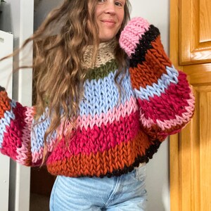 Puede incluir: Un suéter de punto grueso con cuello alto y mangas anchas. El suéter presenta rayas horizontales en tonos de rosa, rojo, naranja, azul y beige. Las mangas tienen un ribete negro con un toque de brillo.