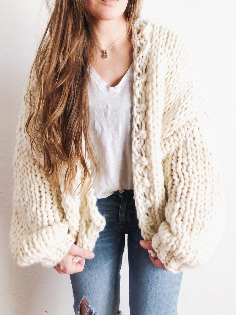 May include: A white chunky knit cardigan with a loose fit and long sleeves. The cardigan is open at the front and has a simple design.