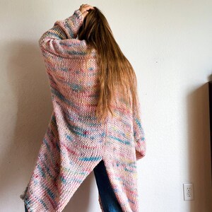 Op de afbeelding: Een lange, roze cardigan met een kleurrijk, marmerpatroon. De cardigan heeft een diepe V-hals en een split in de rug.