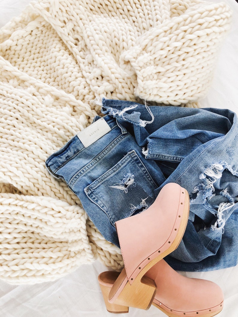 May include: A white chunky knit cardigan, blue denim jeans with distressing, and a pair of pink leather clogs with wooden soles.
