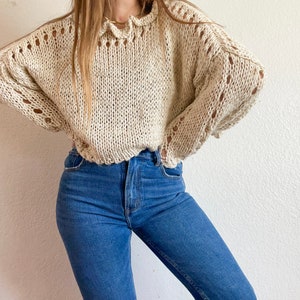 May include: A cream-coloured crocheted sweater with a ruffled neckline and open-stitch detailing. The sweater is worn with blue jeans.