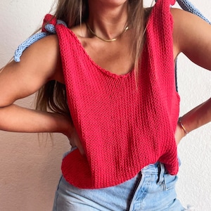 May include: A red knitted tank top with blue straps tied at the shoulders. The tank top is cropped and has a scoop neck. The model is wearing denim shorts.