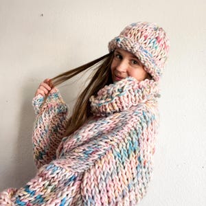 May include: A colorful knitted sweater and matching beanie hat in shades of pink, blue, and yellow. The sweater has a loose, oversized fit and a high neck.