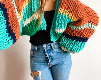 Könnte beinhalten: Ein farbenfroher gestreifter Cardigan mit einer grobmaschigen Strickstruktur. Der Cardigan ist kurz geschnitten und hat lange Ärmel. Die Farben sind eine Mischung aus Orange, Gelb, Grün, Blau und Weiß.
