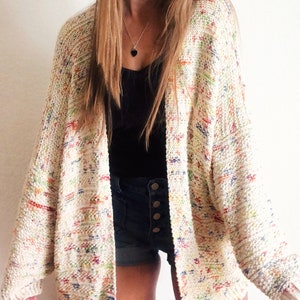 Könnte beinhalten: Ein weißer Cardigan mit einem bunten, gesprenkelten Muster. Der Cardigan hat eine lockere Passform und lange Ärmel.