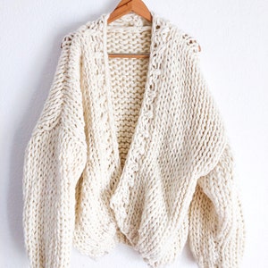 May include: A cream-coloured chunky knit cardigan with a V-neck and long sleeves. The cardigan is hanging on a wooden hanger.