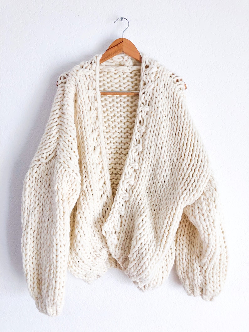 May include: A cream-coloured chunky knit cardigan with a V-neck and long sleeves. The cardigan is hanging on a wooden hanger.