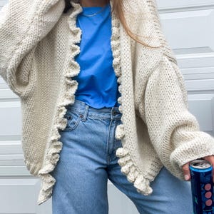 Könnte beinhalten: Cremefarbene, grob gestrickte Strickjacke mit Rüschenkanten. Der offene Pullover wird mit einem blauen T-Shirt und hellblauen Jeans kombiniert. Eine Dose mit blauem und rosa Etikett wird in der Hand gehalten.