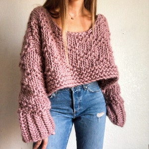 May include: An oversized knitted jumper in a pale pink shade, featuring a loose fit and long sleeves. The jumper has a slightly cropped length and a relaxed, casual style.