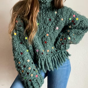 May include: A green knitted sweater with a turtleneck and fringe detailing. The sweater is decorated with iridescent sequins in various colors.