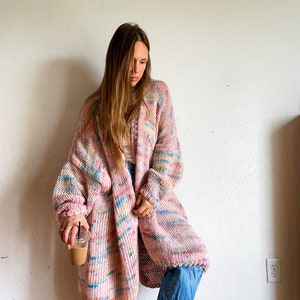 Op de afbeelding: Een lange, roze, blauwe en gele gebreide cardigan met een losse pasvorm. De cardigan is open en de persoon die hem draagt ​​houdt een kop koffie vast.