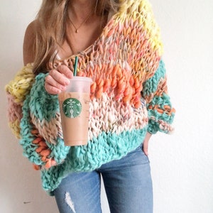May include: A woman wearing a colorful, chunky knit sweater with a teal, pink, and orange color scheme. She is holding a Starbucks iced coffee in a clear plastic cup.