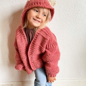 Può includere: Un bambino piccolo indossa un cardigan di maglia rosa con cappuccio e orecchie di maglia bianca. Il cardigan ha un motivo a treccia e si abbottona sul davanti. Il bambino indossa jeans blu.