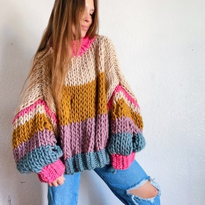 Könnte beinhalten: Ein grob gestrickter Pullover mit horizontalen Streifen in Rosa, Beige, Senfgelb, Lila und Türkis. Der Pullover hat eine lockere Passform und überdimensionale Ärmel.