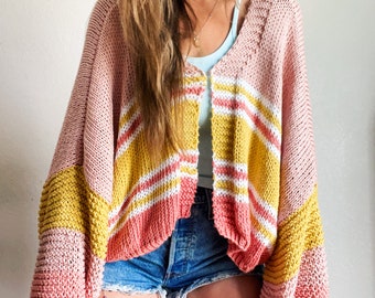 Puede incluir: Un cárdigan abierto de punto en tonos rosa, amarillo y blanco. Las mangas y el cuerpo presentan un diseño de bloques de color con rayas horizontales. El cárdigan se lleva sobre un top azul claro y unos shorts vaqueros.