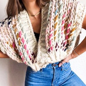 May include: A white knitted cowl with colourful yarn accents. The cowl is worn by a person with long brown hair and is styled with blue jeans.