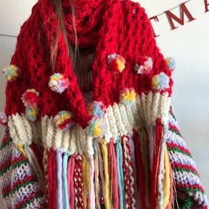 Puede incluir: Una bufanda de punto roja con pompones y flecos de colores. La bufanda está hecha de hilo grueso y tiene un estilo suelto y drapeado.