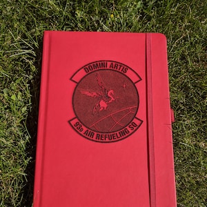 May include: A red notebook with a debossed emblem featuring a winged horse and the text "DOMINI ARTIS" and "93d AIR REFUELING SQ". The notebook has a matching red elastic band closure and a ribbon page marker.