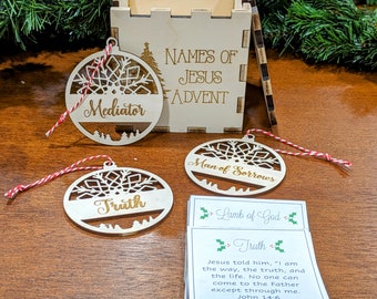 May include: A set of five wooden Christmas ornaments with a snowflake design and gold lettering. The ornaments are in a wooden box with the words "Names of Jesus Advent" engraved on the front. The ornaments feature the names "Mediator", "Truth", "Man of Sorrows", and "Lamb of God".