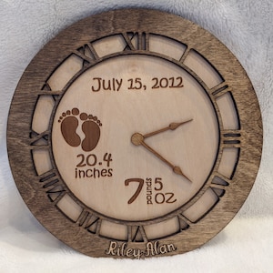 Puede incluir: Un reloj de madera con números romanos y una mancha marrón. La esfera del reloj es de madera clara y presenta un diseño de huella de pie de bebé, la fecha "15 de julio de 2012" y el texto "20,4 pulgadas", "7 libras 5 oz" y "Riley Alan".