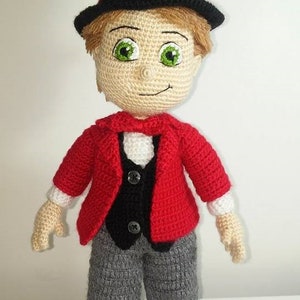 May include: A crocheted amigurumi doll wearing a red jacket, black vest, gray pants, and a black top hat with a red band. The doll has brown hair, a beige face, and green eyes. The doll is standing on a white surface. "JOLLY KIDS AMIGURUMI DESIGNS" is written below the doll.