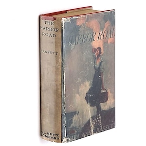 May include: The Harbor Road by Sara Ware Bassett, a vintage book with a cover featuring a woman in a dress standing on a rocky shore with a basket in her hand, looking out at the sea.