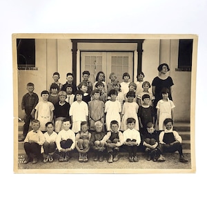 May include: A black and white photo of a group of children, mostly girls, standing in front of a doorway. They are all dressed in clothing from the early 1900s. There is a woman standing in the back, also dressed in clothing from the early 1900s.