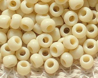May include: A close-up image of a pile of small, round, cream-colored beads. The beads are made of plastic and have a smooth, glossy finish.