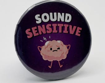 May include: A round, purple button with the words "SOUND SENSITIVE" in white and pink. A cartoon brain with a sad expression is in the center. The button is a fun accessory.