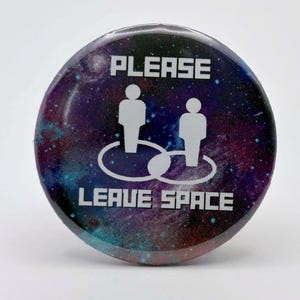 May include: Round button with a galaxy design in shades of purple, blue, and teal. The button features the text "PLEASE LEAVE SPACE" in white, along with two white silhouette figures within a white circle.