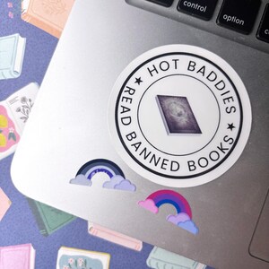 May include: A circular white sticker with black text reading HOT BADDIES READ BANNED BOOKS and a central illustration of a book, accompanied by two smaller rainbow stickers with clouds in shades of blue, purple, and pink on a laptop surface.