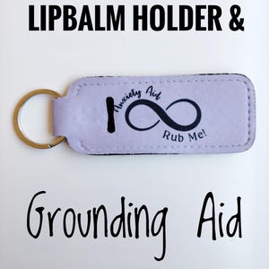 May include: A lavender lip balm holder and grounding aid keychain with a gold-colored ring. The holder has the text "Anxiety Aid" and "Rub Me!" with an infinity symbol. The words "Lipbalm Holder & Grounding Aid" are also displayed.